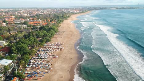 Aerial-flyover-sandy-Legian-Beach-with-beautiful-landscape-during-sunlight,-waves-crashing-on-shore-and-many-sunshades