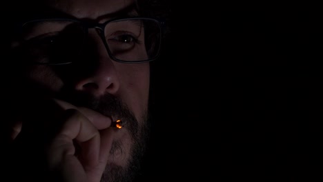 Young-adult-male-with-glasses-and-beard-smoking-cigarette-in-dark-room-reflecting-digital-screen