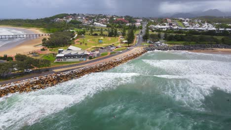 People-Surfing-And-Swimming-At-Coffs-Harbour-Near-Solitary-Islands-Marine-Park---Vehicles-Driving-On-Seaside-Road-With-Coffs-Harbour-Jetty-In-NSW,-Australia