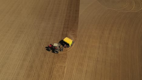 Tracking-aerial-view-of-a-red-double-wheeled-tractor-sowing-seeds-on-a-agricultural-field-with-a-yellow-seeder-during-spring-time