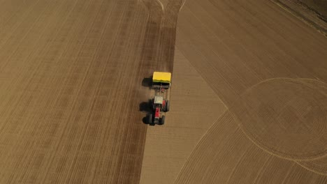 Tracking-aerial-view-of-a-red-double-wheeled-tractor-sowing-seeds-in-a-straight-line-on-a-agricultural-field-with-a-yellow-seeder-during-spring-time