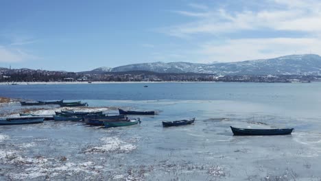 Aerial-shot-of-the-half-frozen-lake-and-boats-docked-on-the-shore-on-sunny-day-with-city-and-mountains-the-in-the-background