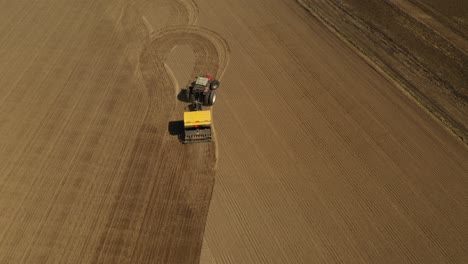 Tracking-aerial-view-of-a-red-double-wheeled-tractor-sowing-seeds-on-a-agricultural-field-with-a-yellow-seeder-during-spring-time