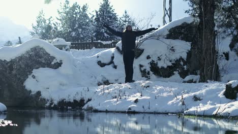 Woman-standing-next-to-a-lake-during-winter,-opening-arms-wide-to-the-side