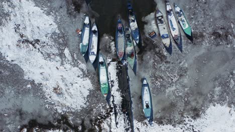 Aerial-view-of-small,-colourful-boats-docked-in-a-half-frozen-lake-during-wintertime