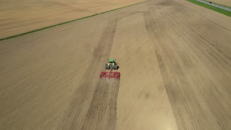 Tracking-aerial-view-of-green-double-wheeled-tractor-cultivating-agricultural-field-with-red-cultivator-during-spring-time