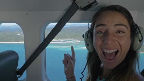 Cheerful-And-Excited-Girl-Wearing-Headphones-Pointing-To-The-Beach-Seen-Through-Window-Of-Private-Plane