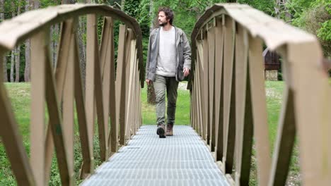 Young-man-in-his-20s-wearing-neutral-clothing-walking-over-a-small-footbridge-towards-the-camera-on-an-outdoors-hiking-trail-in-Catalonia
