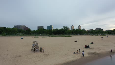 aerial-video-zooming-out-of-monrose-beach-in-chicago,-showing-people-playing-on-the-beach-and-the-city-in-the-background