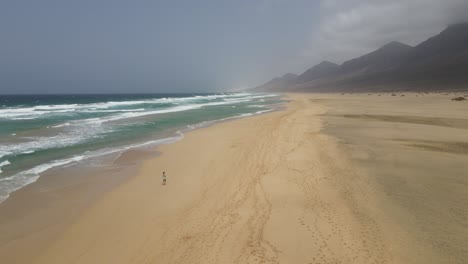 aerial-drone-shot-of-a-girl-running-on-a-wide-white-beach-with-big-waves-crashing-and-mountains-in-the-background-in-Fuerteventura,-canary-island
