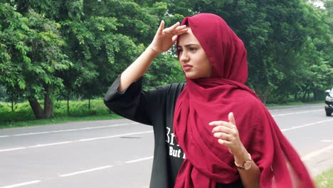 Slow-motion-of-a-young-smiling-Afghan-girl-in-hijab-in-a-black-and-red-dress-standing-and-hiding-face-from-sun-on-the-side-of-the-road-waiting-for-public-transport-to-go-back-home