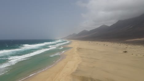 Toma-Aérea-De-Grandes-Olas-Rompiendo-En-La-Larga-Playa-Blanca-De-Cofete-Con-Hermosas-Montañas-En-El-Fondo-En-Fuerteventura,-Isla-Canaria