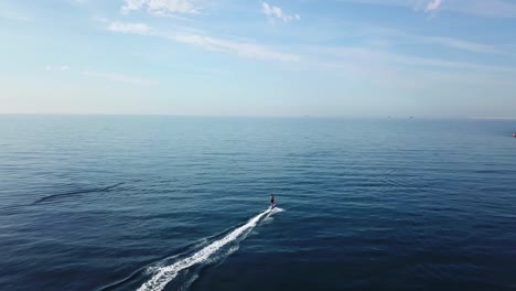 Surfer-riding-a-fast-electric-surfboard-into-the-open-sea-on-a-calm-sunny-day,-aerial-follow