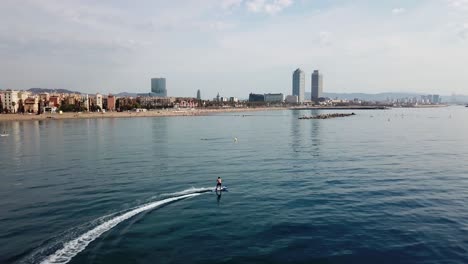 Aerial-of-surfer-riding-electric-surfboard-with-great-views-over-the-skyline-of-Barcelona,-Spain