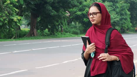 Beautiful-serious-young-Afghan-woman-in-hijab-holding-file-and-bag-in-hand-and-standing-on-street-near-road,-charming-female-in-black-t-shirt
