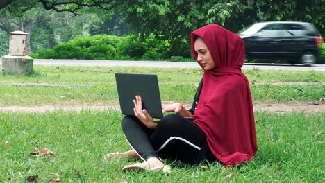 Glückliche-Afghanische-Frau-Im-Hijab,-Die-An-Einem-Laptop-Auf-Grünem-Rasen-In-Der-Nähe-Einer-Stark-Befahrenen-Straße-Arbeitet