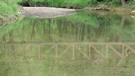 Reflexion-Im-Fluss-Entlang-Eines-Naturpfades-Eines-Mannes,-Der-über-Eine-Kleine-Holzbrücke-Geht