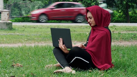 Young-afghan-female-student-sits-on-the-ground-in-a-park-near-busy-road-on-a-sunny-day,-studies-using-a-computer-and-does-homework