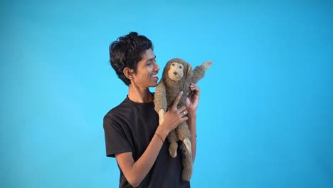 Fun-young-Indian-boy-in-basic-black-t-shirt-isolated-on-blue-background-studio-and-playing-with-toy-monkey