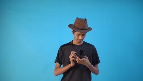 Indischer-Jungenfotograf-überprüft-Das-Neue-Kameragehäuse-In-Schwarzem-T-shirt-Und-Blauem-Innenstudiohintergrund-Und-Trägt-Braunen-Sheriffhut