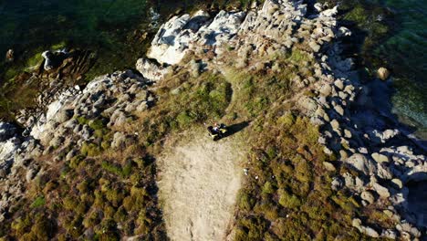 Aerial-view-of-ATV-drives-to-the-edge-of-a-land-covered-in-rocks-and-bushes-with-blue-sea-in-front