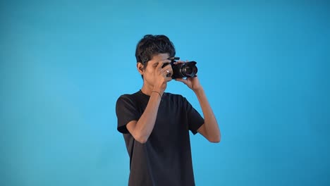 El-Joven-Fotógrafo-Indio-Está-Tomando-Algunas-Imágenes-Fijas-Con-Su-Cámara-Dslr-En-Una-Camiseta-Negra-Y-Un-Fondo-De-Estudio-Interior-De-Color-Azul