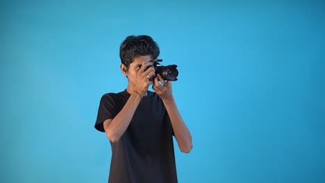 Hübscher-Junger-Fotograf,-Der-Die-Kamera-Untersucht-Und-Den-Fokus-Im-Blauen-Hintergrund-Des-Fotostudios-Anpasst