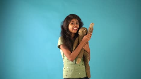 A-young-Indian-girl-in-green-t-shirt-playing-with-a-monkey-doll-saying-'Hi'-seeing-the-camera-standing-in-an-isolated-blue-background-studio