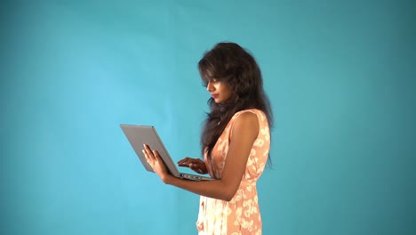 Side-view-of-a-young-Indian-girl-in-orange-frock-with-laptop-standing-in-an-isolated-blue-background