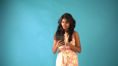 A-young-Indian-girl-in-orange-frock-wearing-earphones-and-talking-in-video-chat-with-phone-standing-in-an-isolated-blue-background