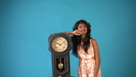 A-young-Indian-girl-in-orange-frock-thinking-in-front-of-an-old-vintage-clock-in-an-isolated-blue-background