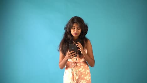 A-young-Indian-girl-in-orange-frock-wearing-earphones-and-talking-in-the-video-chat-with-phone-standing-in-an-isolated-blue-background