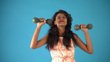 A-young-Indian-girl-in-orange-frock-lifting-dumbbells-smiling-at-the-camera-standing-in-an-isolated-blue-background