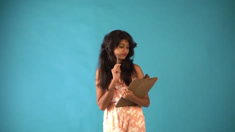 A-young-Indian-girl-in-orange-frock-with-a-notepad-and-pencil-thinking-and-standing-in-an-isolated-blue-background