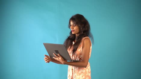 A-young-Indian-girl-in-orange-frock-doing-video-chat-in-laptop-and-standing-in-an-isolated-blue-background