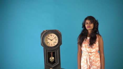 A-young-Indian-girl-in-orange-frock-standing-and-showing-the-old-vintage-clock-in-an-isolated-blue-background