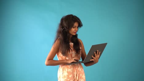 Side-view-of-a-young-Indian-girl-in-orange-frock-working-with-a-laptop-in-hand-standing-in-an-isolated-blue-background