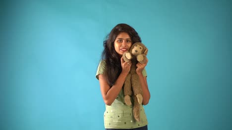 A-cute-young-Indian-girl-in-green-t-shirt-playing-with-a-monkey-doll-seeing-the-camera-standing-in-an-isolated-blue-background-studio