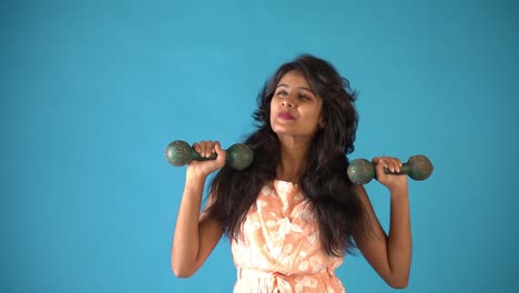 A-young-Indian-girl-in-orange-frock-lifting-dumbbells-one-by-one-smiling-at-the-camera-standing-in-an-isolated-blue-background