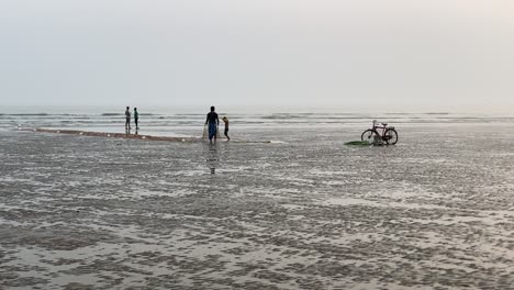 Silhouette-view-of-a-beach-with-waves,-people-taking-net-for-fishing-in-front-of-cycle-and-some-people-talking-in-a-empty-beach-in-India