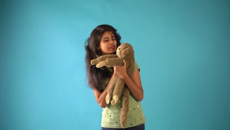 A-young-Indian-girl-in-green-t-shirt-cuddling-with-a-monkey-doll-standing-in-an-isolated-blue-background-studio