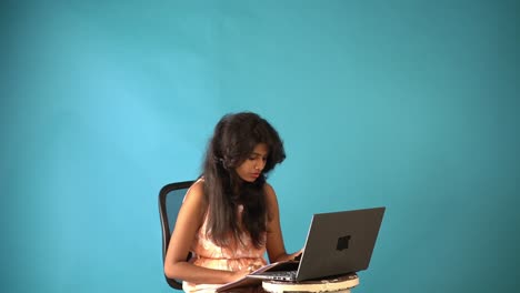 A-young-Indian-girl-in-orange-frock-sitting-in-front-of-laptop-and-writing-exam-in-notepad-sitting-in-an-isolated-blue-indoor-background