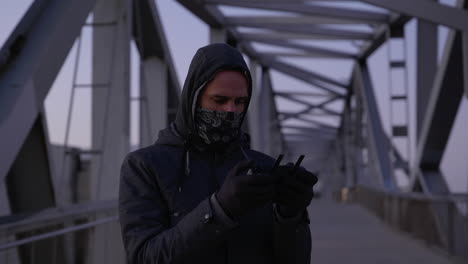 Mysterious-masked-hooded-male-watching-drone-controller-flying-from-iron-girder-bridge