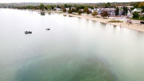 Drone-shot-flying-over-Crystal-Lake-and-the-small-town-of-Beulah-Michigan-during-autumn-with-foliage-changing-showcasing-clear-water,-beautiful-beach-and-homes-with-mountains-in-background