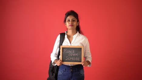 A-young-Indian-girl-in-white-shirt-carrying-a-slate-written-'please-open-school'-looking-the-camera-standing-in-an-isolated-red-background-studio