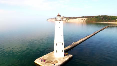 Drone-shot-flying-away-from-a-beautiful-white-lighthouse-on-Lake-Michigan-with-cliffs-in-the-background-and-people-walking-along-the-pier-to-the-lighthouse