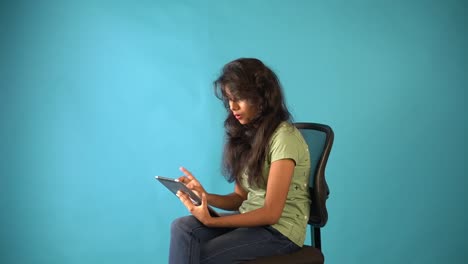 A-young-Indian-girl-in-green-t-shirt-talking-in-video-chat-in-tab-sitting-on-a-chair-in-an-isolated-blue-background-studio