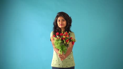 A-young-Indian-girl-in-green-t-shirt-showing-her-bouquet-of-flowers-looking-the-camera-with-shy-expression-standing-in-an-isolated-blue-background-studio