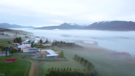Ein-Lkw-Panning-Eines-Fjords-Sehr-Früh-Am-Morgen-Mit-Gebäuden-Und-Viel-Nebel-Und-Dunst-Mit-Schneebedeckten-Bergen-Im-Hintergrund-Und-Hellgrünen-Bäumen-Im-Vordergrund