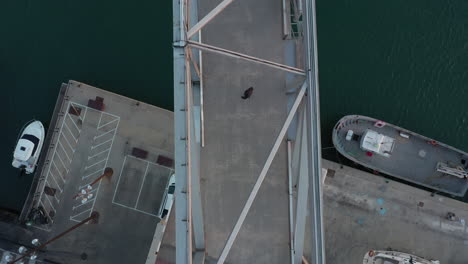 Birdseye-aerial-view-above-male-walking-across-bridge-looking-behind-and-begins-to-run-scared
